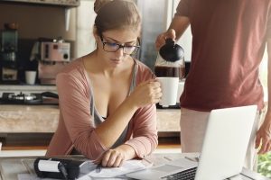 worried-young-woman-calculating-family-expenses-doing-domestic-budget-using-generic-laptop-calculator-kitchen-while-her-husband-standing-her-pouring-hot-coffee-into-her-mug_273609-1688